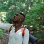 Fear Failure - African American male with backpack standing in forest and holding mobile phone while catching GPS signal during hike