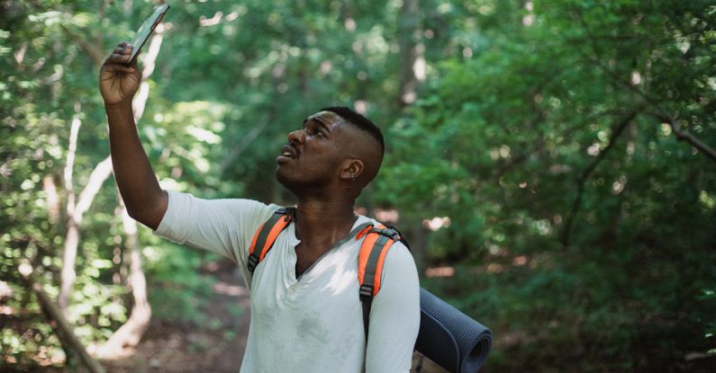 Fear Failure - African American male with backpack standing in forest and holding mobile phone while catching GPS signal during hike