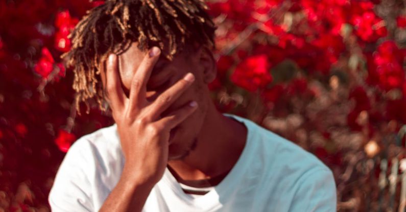 Failure Growth - Annoyed African American male covering face with hand while sitting in park with bright flowers on tree branches