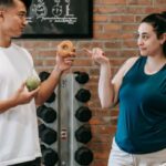 Nutrition Mental - Side view of Asian personal instructor with apple and doughnut offering healthy food to plus size woman while standing in gym