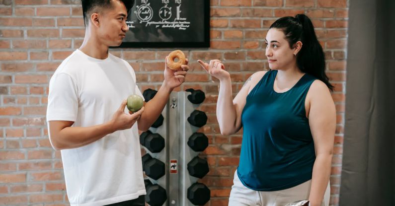 Nutrition Mental - Side view of Asian personal instructor with apple and doughnut offering healthy food to plus size woman while standing in gym