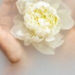Nature Therapy - Unrecognizable female with soft manicured hands holding white flower with delicate petals in hands during spa procedures